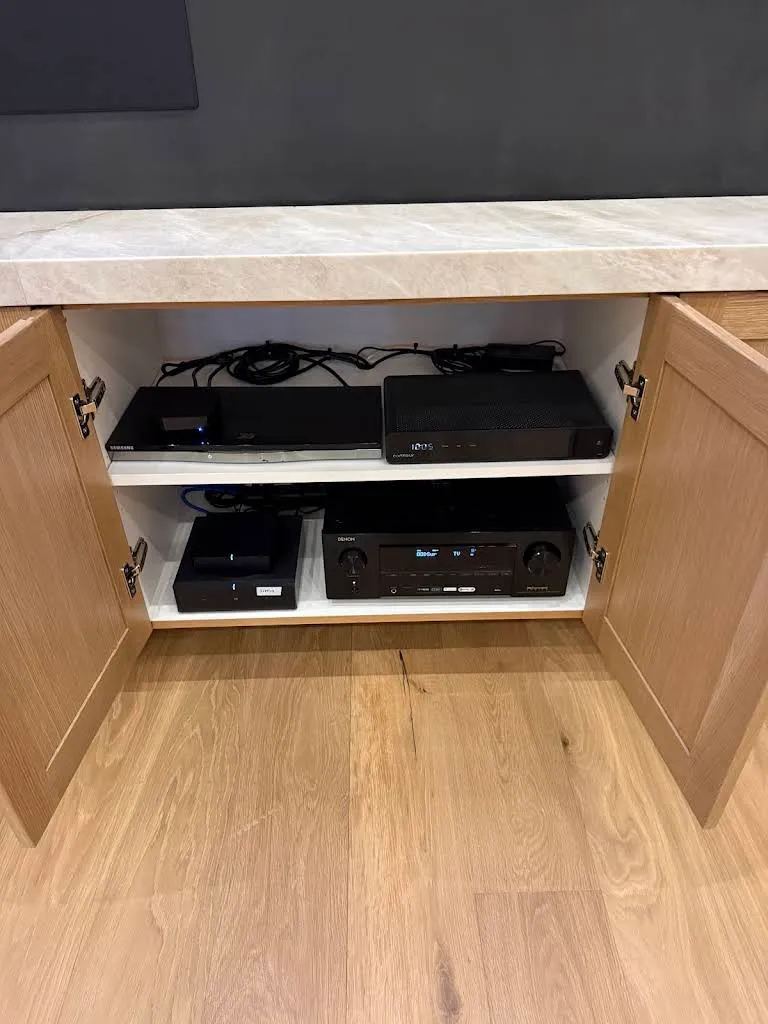 Open media cabinet under a TV showing a Denon AV receiver, a Sonos streamer, and a Blu-ray player on floating shelves above white oak flooring.