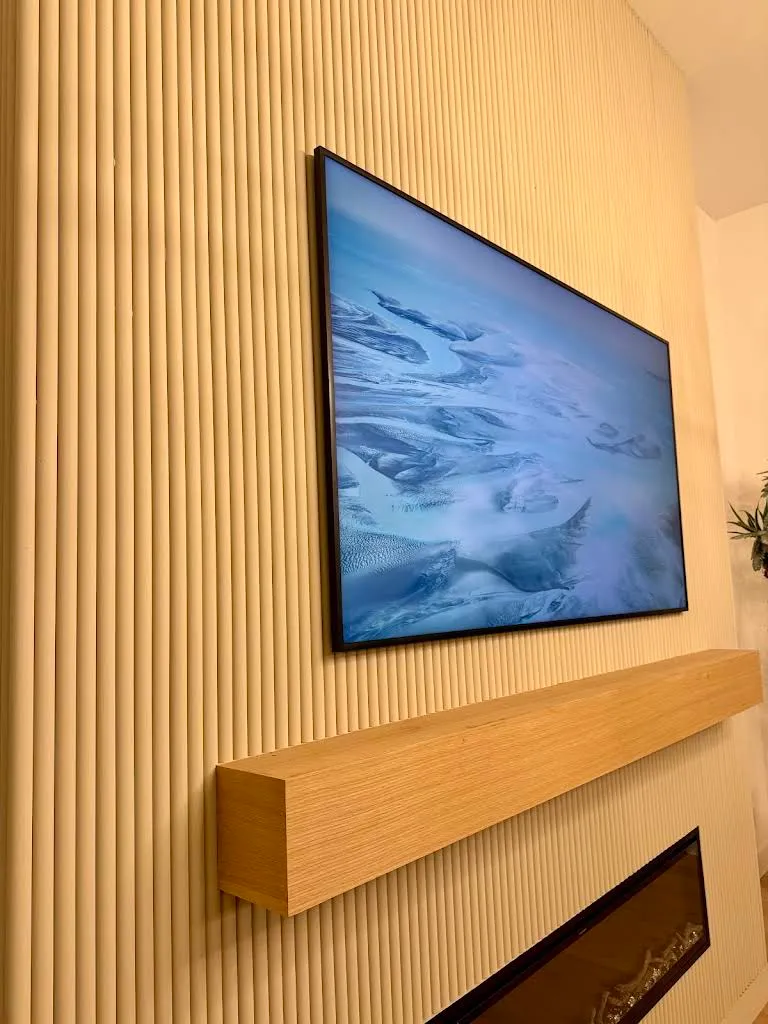 Framed art-mode TV displaying a glacier photograph, mounted on a fluted wood-slat wall above a floating white oak mantel and a linear fireplace.