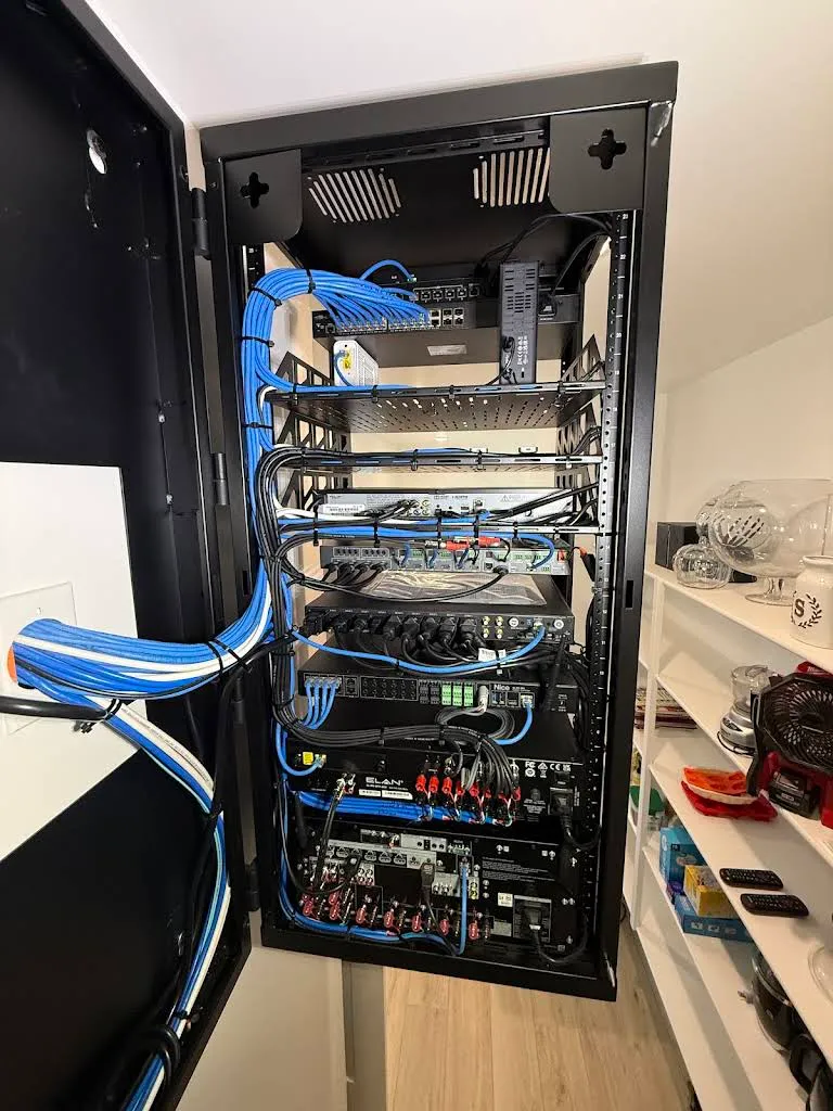 Open in-wall structured wiring rack with a control system, amplifiers, and blue Cat cabling bundled and combed along the side.