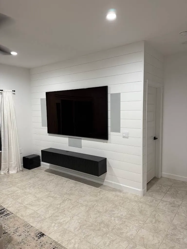 Large TV wall-mounted on a white shiplap accent wall, flanked by flush in-wall speakers with a center channel and a floor subwoofer below.