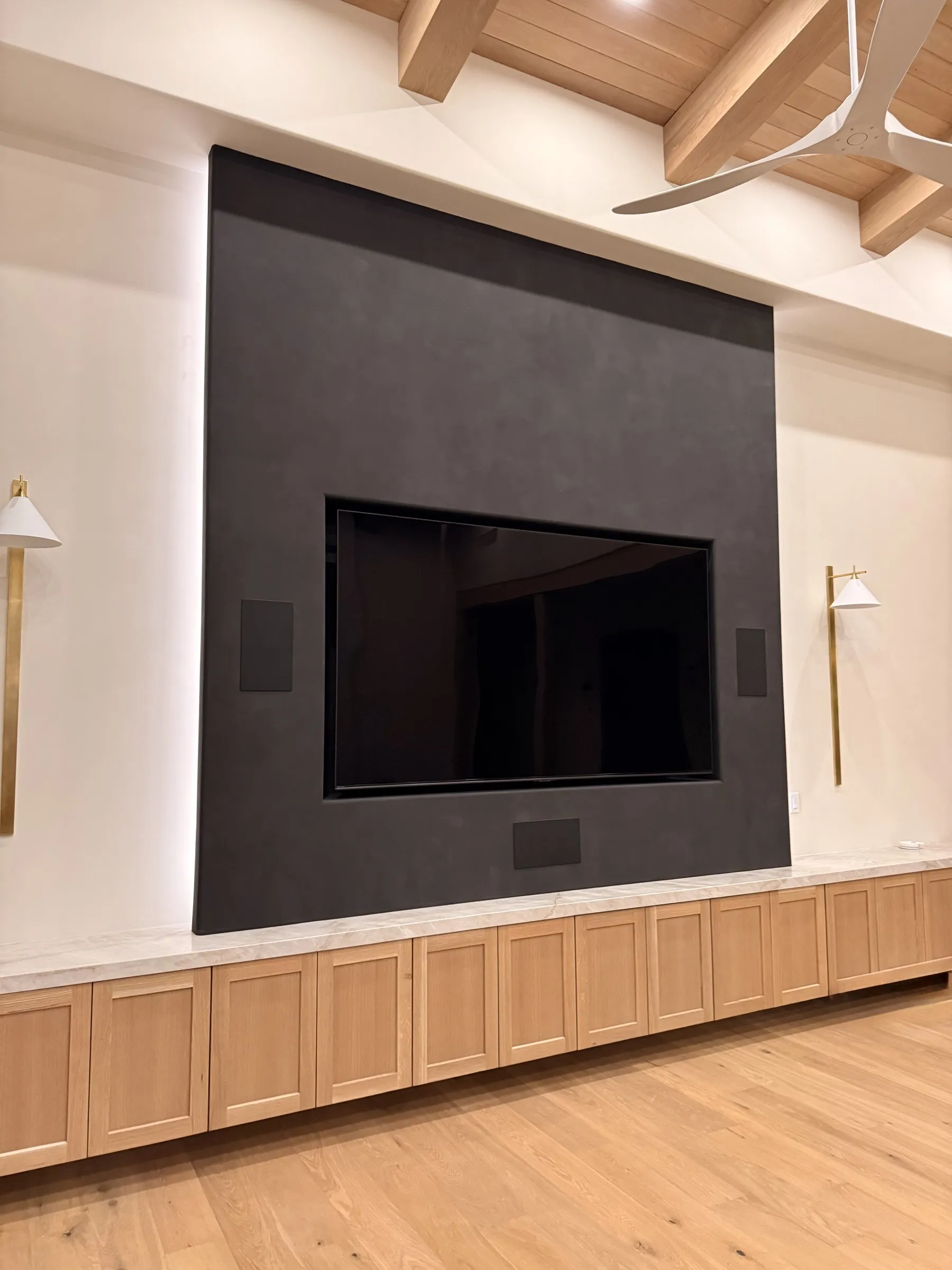 Large TV mounted on a dark stone accent wall above a light wood built-in cabinet, flanked by brass sconces, under an exposed wood-beam ceiling with a fan.