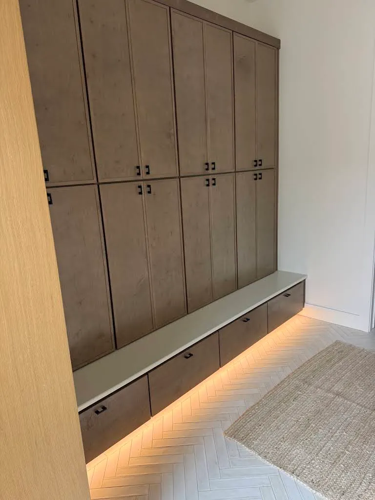 Custom mudroom cabinetry with warm LED accent lighting glowing beneath the bench and toe-kick, over a herringbone tile floor.