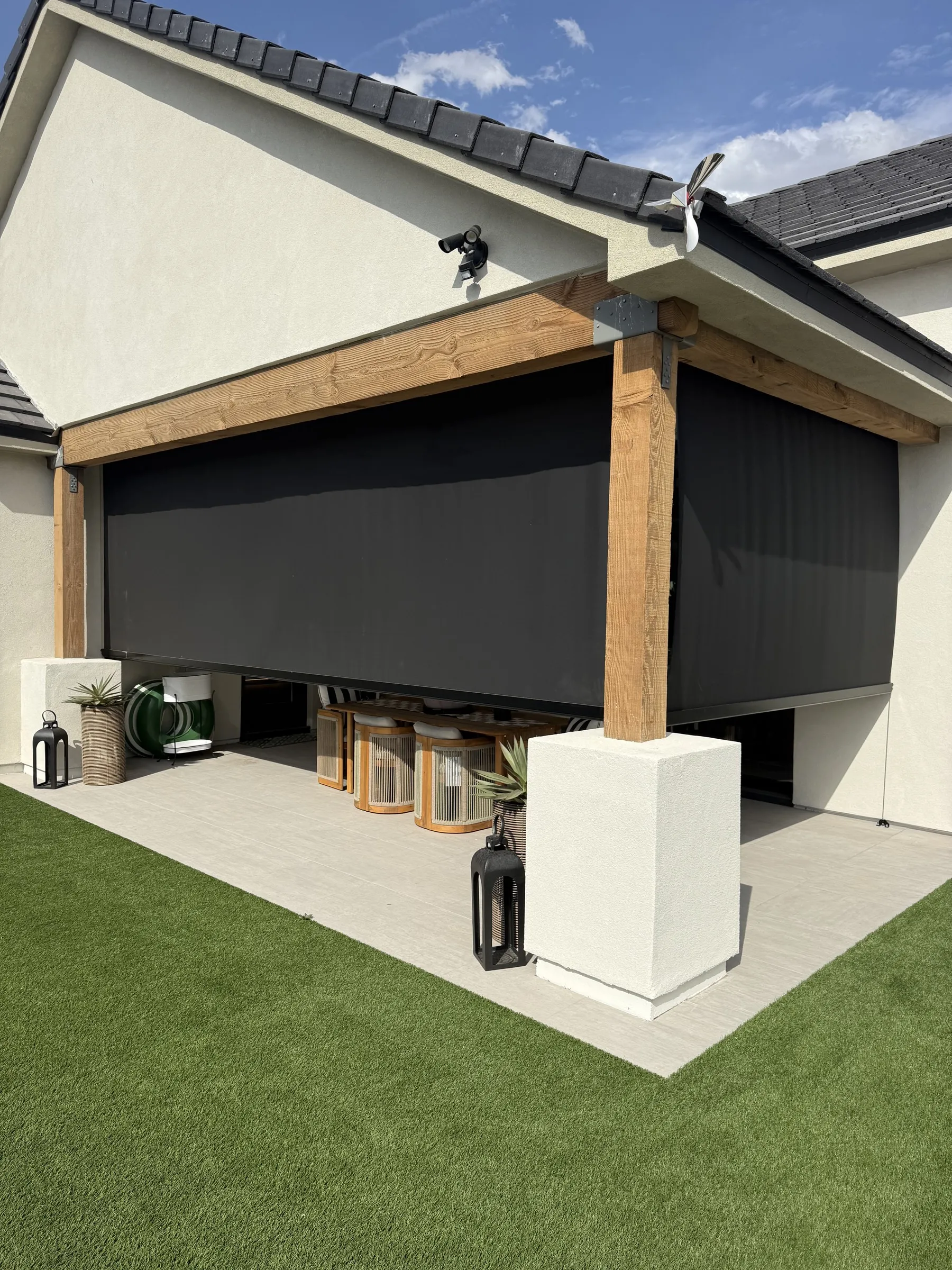 Covered patio pergola with a motorized black solar shade lowered between wood beams, overlooking a turf backyard with potted plants and lanterns.