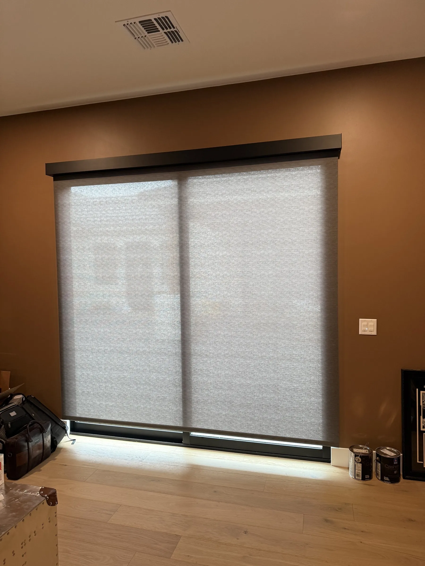 Close-up of a motorized dual roller shade lowered over a sliding glass door in a room with warm accent-lit walls and wood flooring.