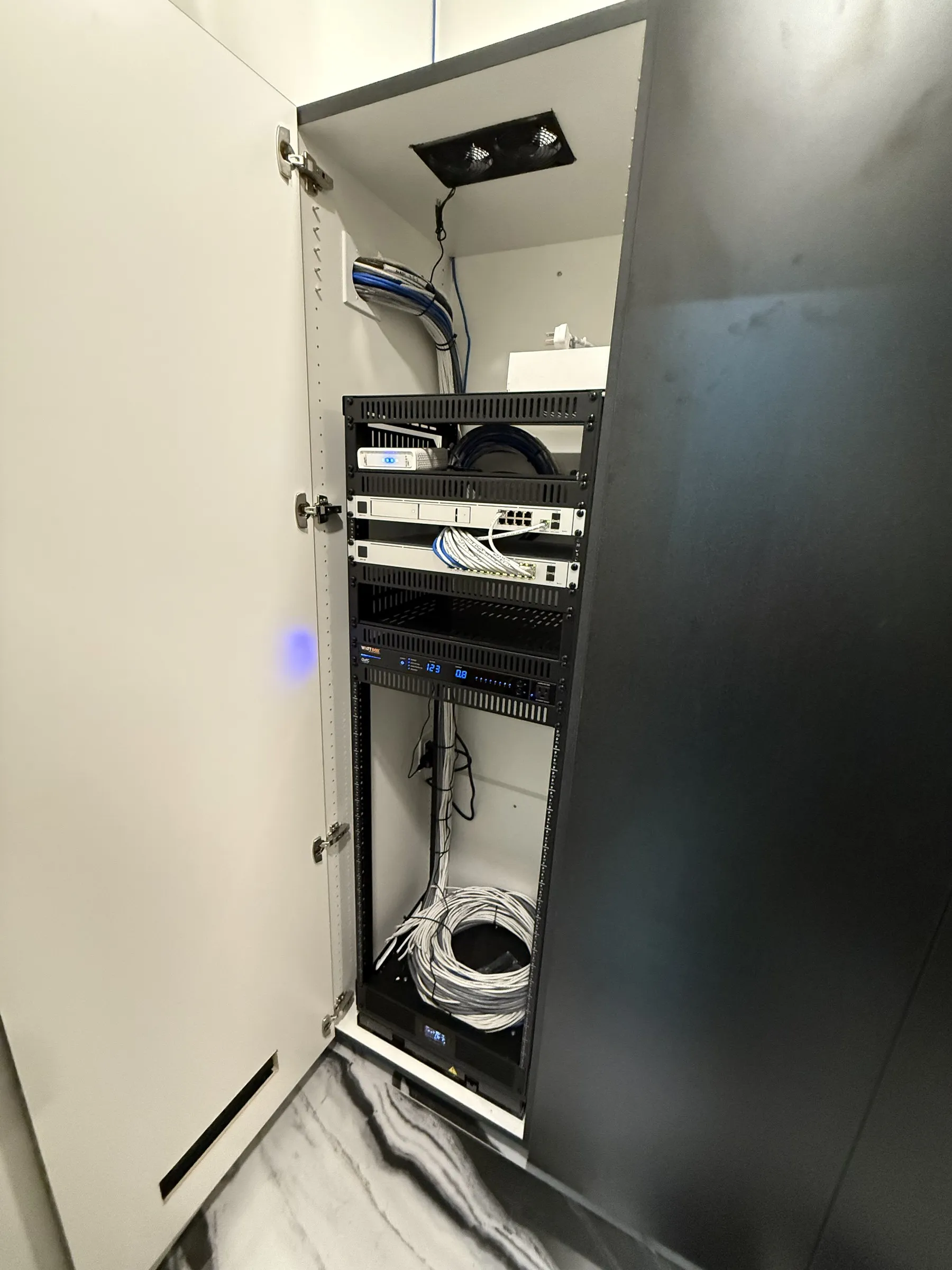 Open equipment rack installed inside a white in-wall cabinet, with network switches, a patch panel, and neatly routed cabling.