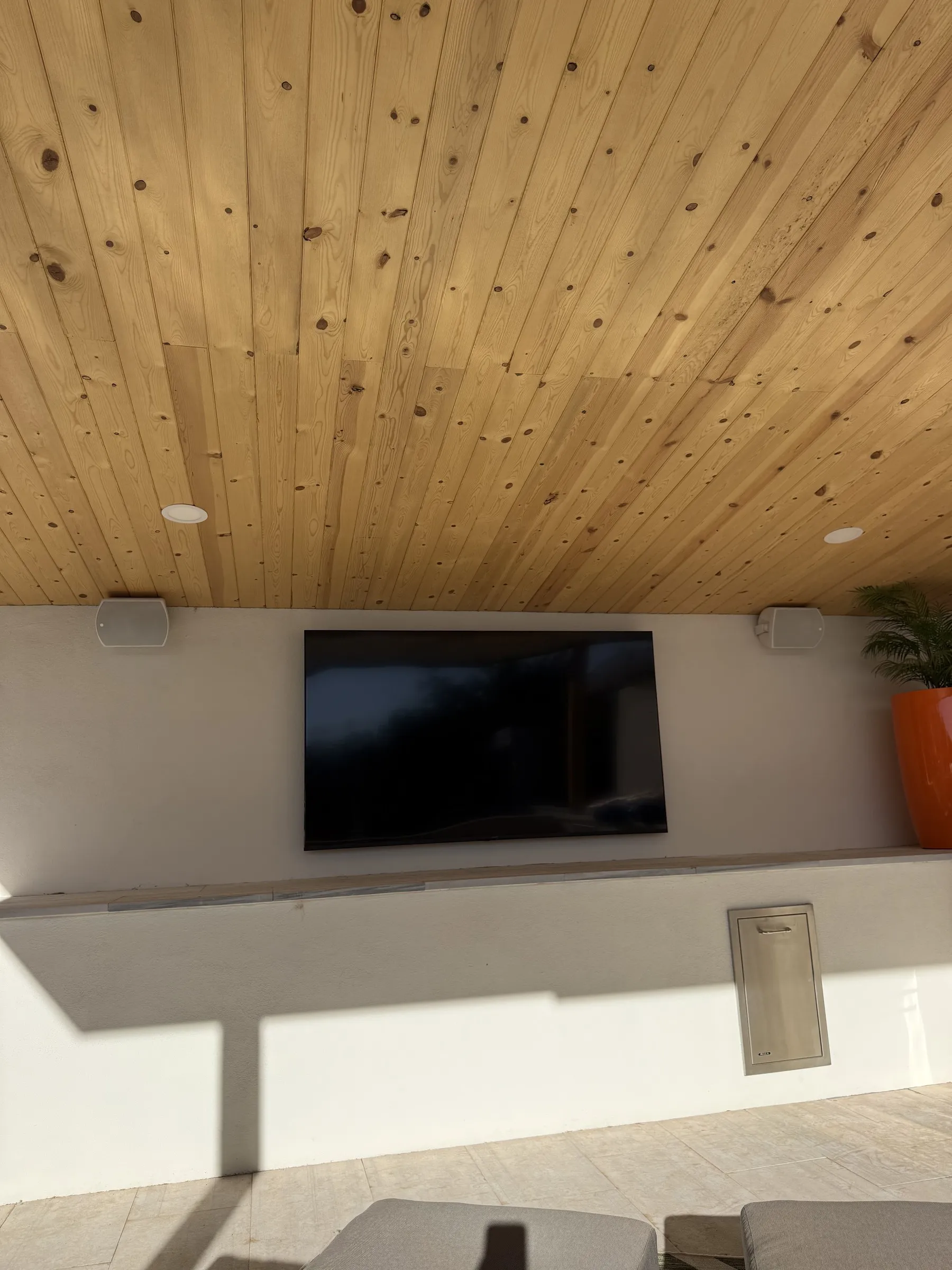 Wall-mounted TV on a covered patio with a wood-plank ceiling, speakers flanking the display, and lounge seating below.