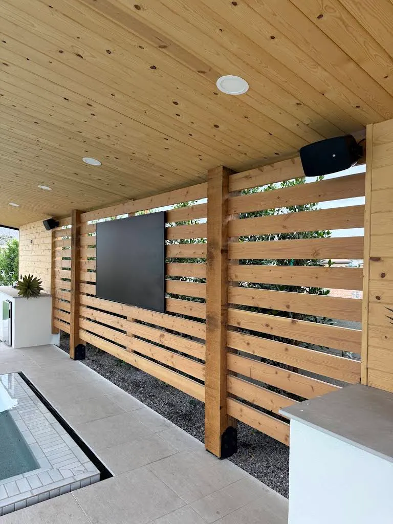 Weatherproof outdoor TV mounted on a cedar-slat privacy wall beneath a wood patio ceiling, with a black JBL outdoor speaker overhead beside a pool.