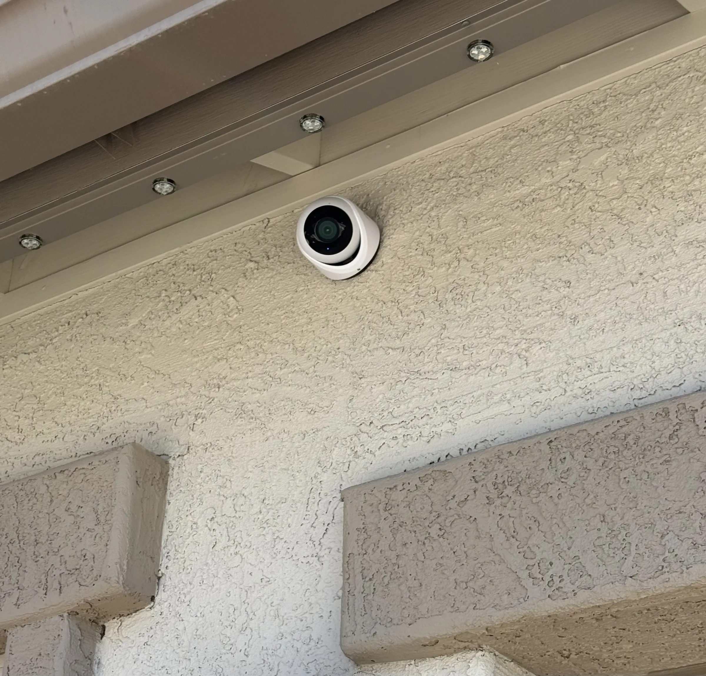 Close-up of a white security camera mounted under a metal eave against a stucco wall, mounting screws visible.