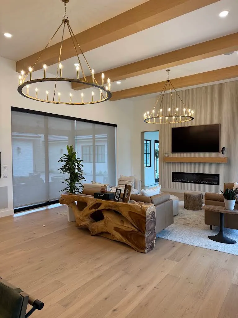 Modern Arizona great room with two large circular chandeliers, a wall-mounted TV above a linear fireplace on a wood-slat wall, and motorized solar shades lowered over floor-to-ceiling windows.