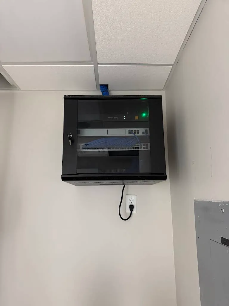 Wall-mounted lockable network cabinet housing a WattBox and a network switch with blue cabling, installed beside an electrical panel under a drop ceiling.